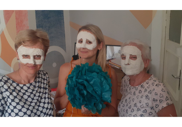 Trzy kobiety pozujące w maskach gipsowych. Kobieta po środku trzyma kwiat zrobiony z bibuły.