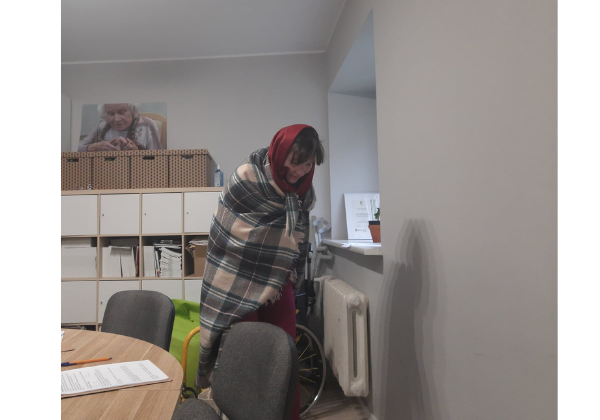 Kobieta z chustą na głowie i szalu wciela się w rolę uchodźcy. Znajduje się w biurze.