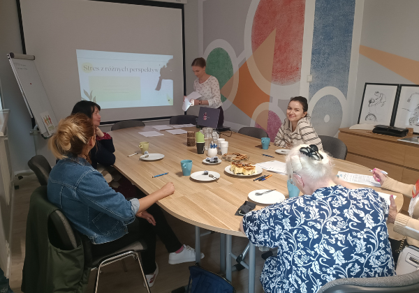 Grupa uśmiechniętych osób siedzących przy stole. Na tablicy jest wyświetlany tytuł Wolontariat a stres. Obok stoi prelegentka prowadząca warsztaty.