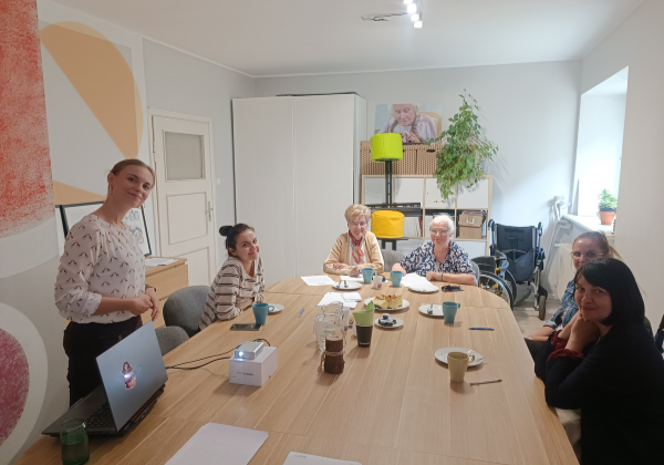 Grupa uśmiechniętych osób siedzi przy stole w biurze. Obok nich stoi prelegentka prowadząca warsztaty.