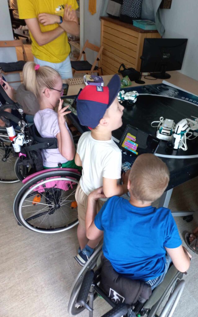 Dzieci przyglądają się robotom rozłożonym na macie. Większość dzieci porusza się na wózku.