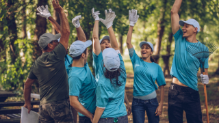 Grupa osób w błękitnych koszulkach i rękawiczkach ogrodniczych wydaję okrzyk radości. Znajdują się w ogrodzie. Pracują w ramach wolontariatu pracowniczego.
