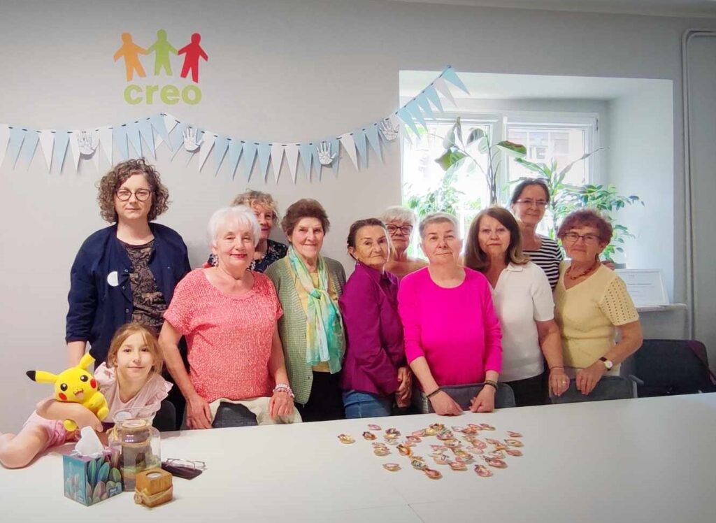 Trzy zdjęcia z warsztatów rękodzielniczych.
Grupa kobiet w sali warsztatowej. 9 kobiet w wieku senioralnym, młoda kobieta i dziewczynka. Przed nimi stół warsztatowy na stole leżą muszelki.