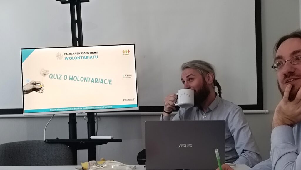 Mężczyzna w sali warsztatowej. Pije kawę. Siedzi przy stole z laptopem przed sobą. Obok niego stoi monitor na stojaku. Na ekranie napis: Qioz o wolontariacie