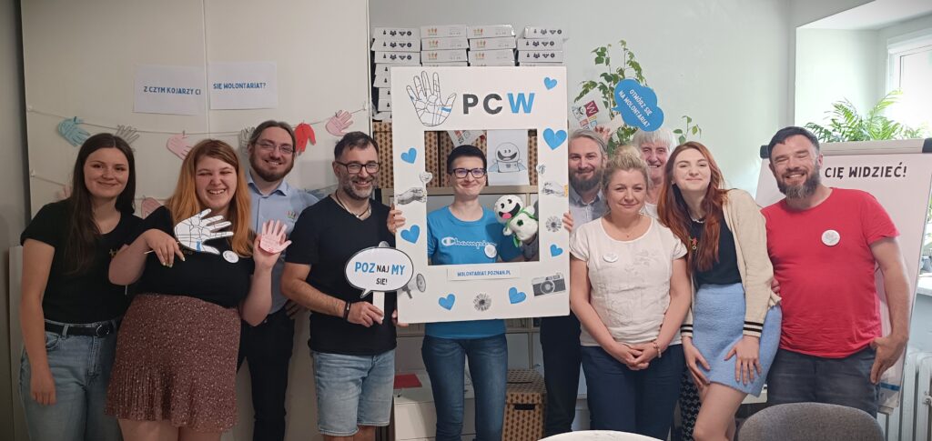 Grupa osób w sali warsztatowej. Stoją i pozują do zdjęcia. Kobieta w centralnej części fotografii trzyma ramkę z