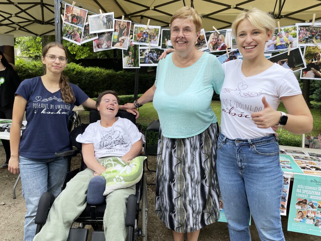 Grupa kobiet pozuje i uśmiecha się do zdjęcia. Jedna z kobiet jest na wózku. Za nimi są powieszone zdjęcia.