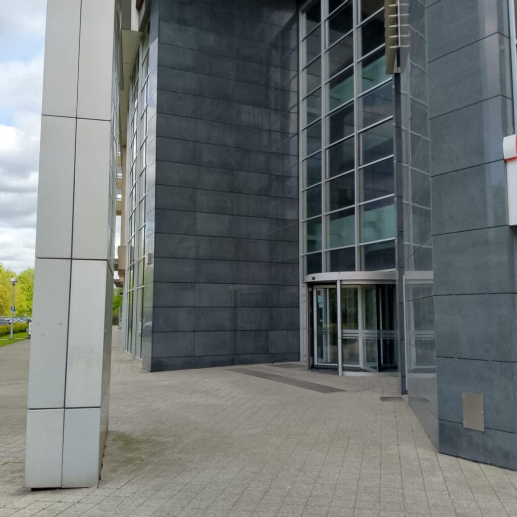 Szary nowoczesny budynek. Z lewej strony kolumna. Z prawej strony przeszklona ściana z drzwiami obrotowymi