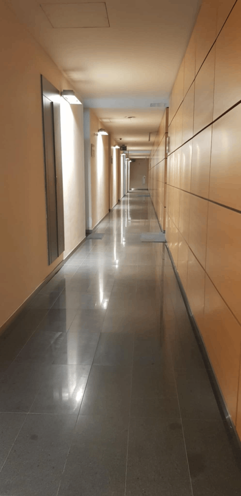 Korytarz podświetlony z lewej strony. Ściany z bloków drewna w jasnym kolorze. Podłoga szara odbijająca światło 