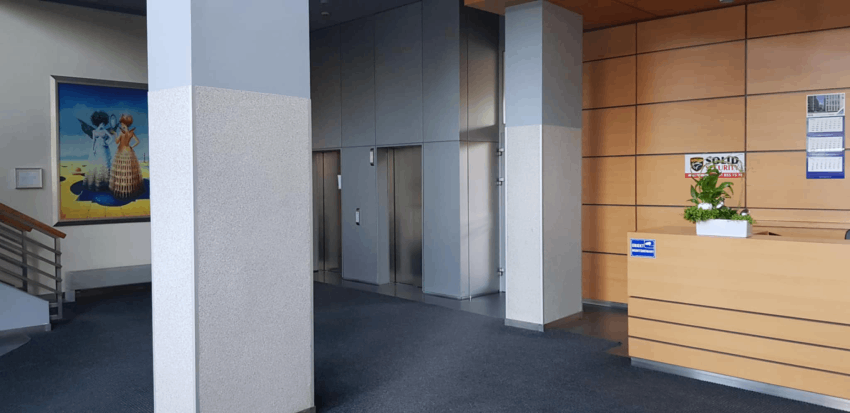 Portiernia na drewniana portiernia, z lewej strony szara prostokątna kolumna, po lewej stronie od niej schody do góry, po prawej dwie windy.