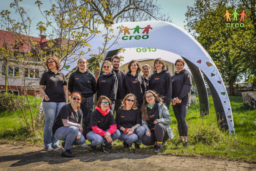 Grupa osób pozuje w plenerze. W tle roślinność i naiot reklamowy CREO