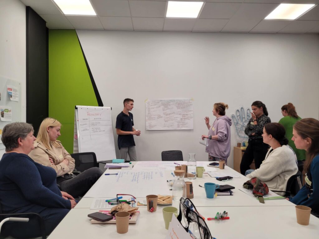 Młody chłopak i dziewczyny prezentują swój projekt. Pozostałe osoby siedzą przy stole i ich słuchają.